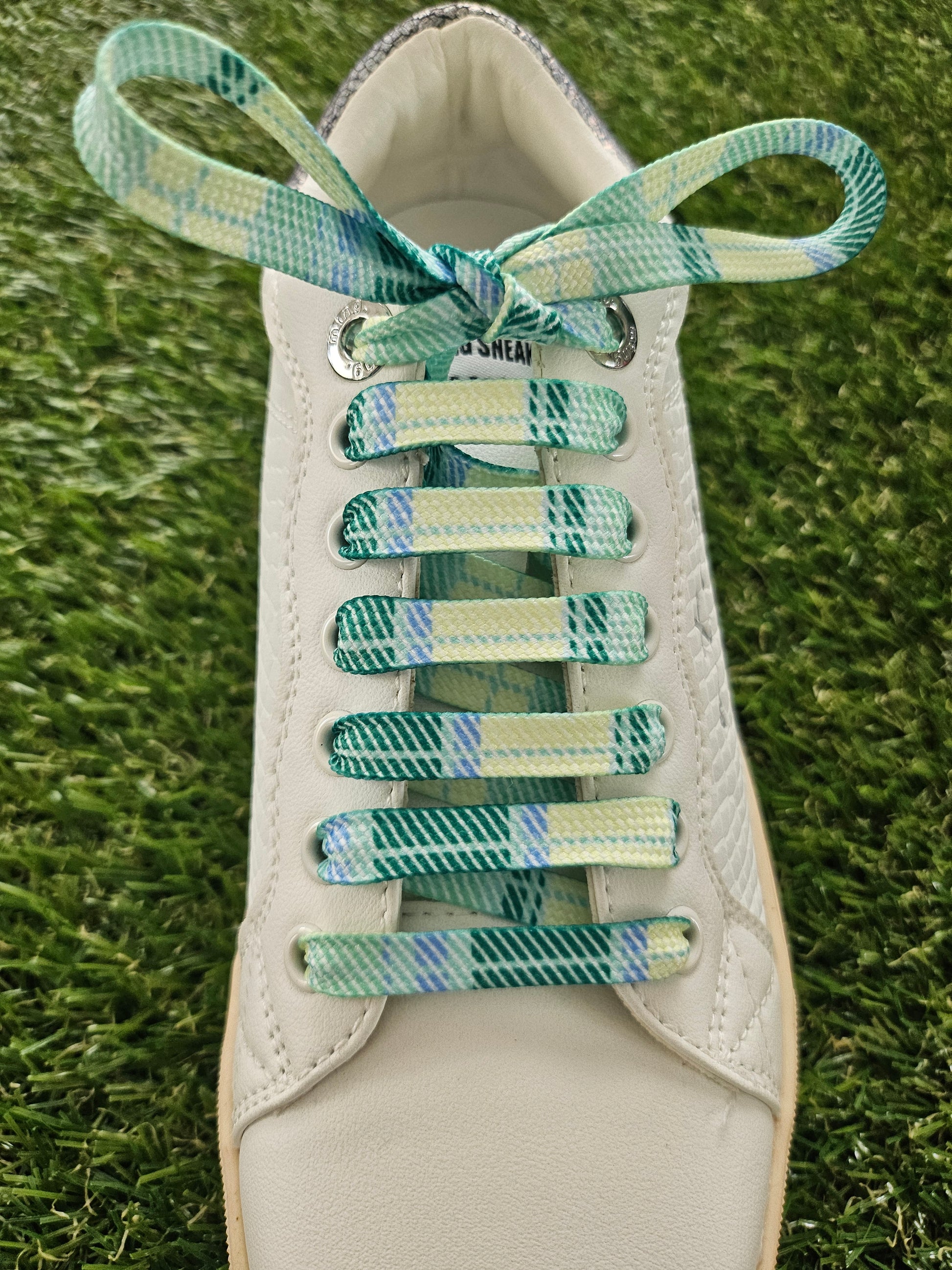 Close-up of a white trainer with green and blue tartan patterned laces on grass