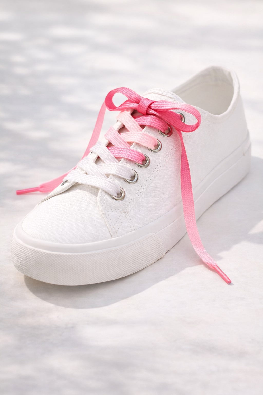 A white training shoe with a shoe lace that has a gradient colour scheme going from white to pink. 