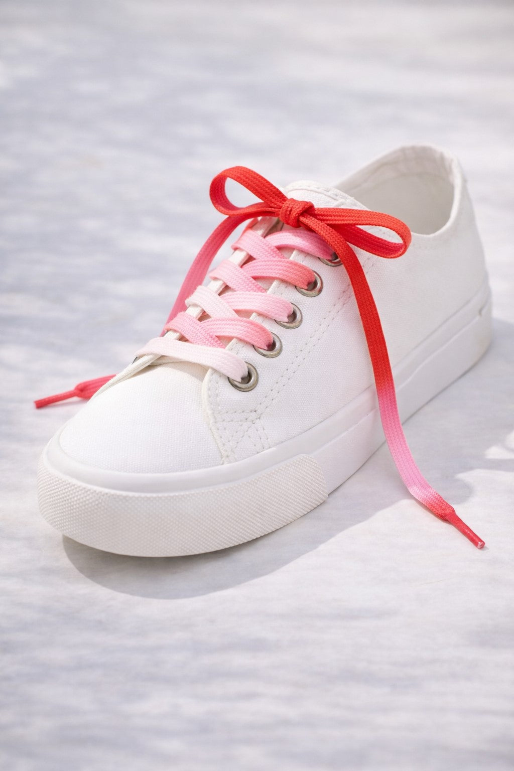 A white training shoe with a shoe lace that has a gradient colour scheme going from white to red. 