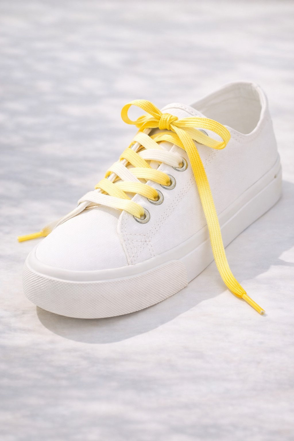 A white training shoe with a shoe lace that has a gradient colour scheme going from white to yellow. 
