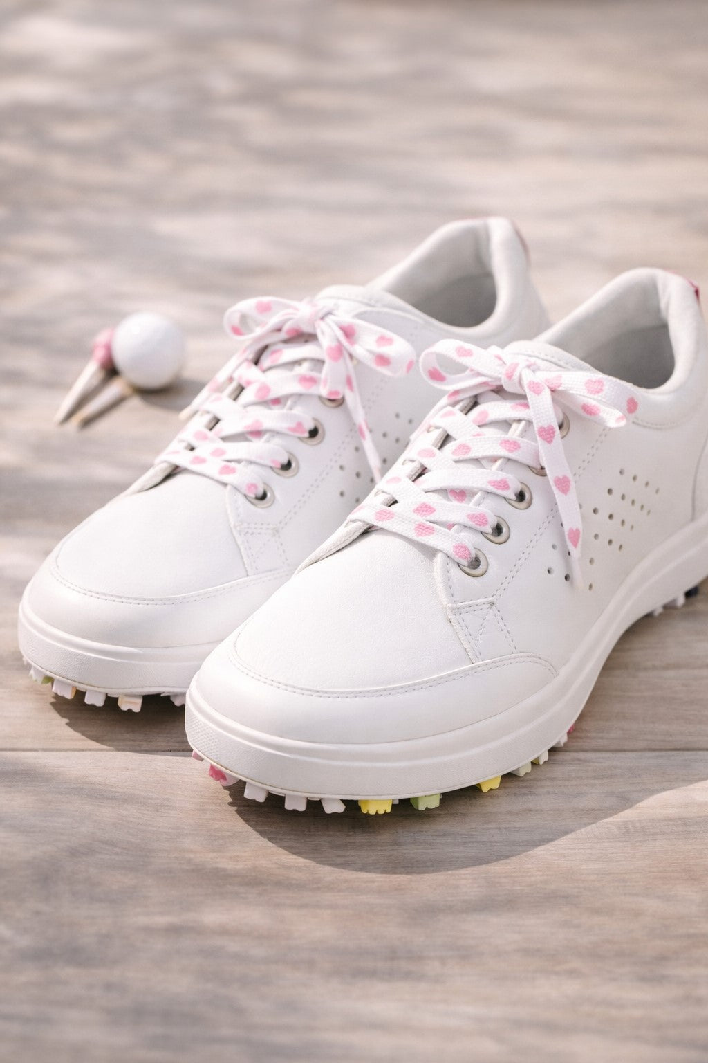 White golf shoes with pink heart laces on a wooden surface with a golf ball and club.