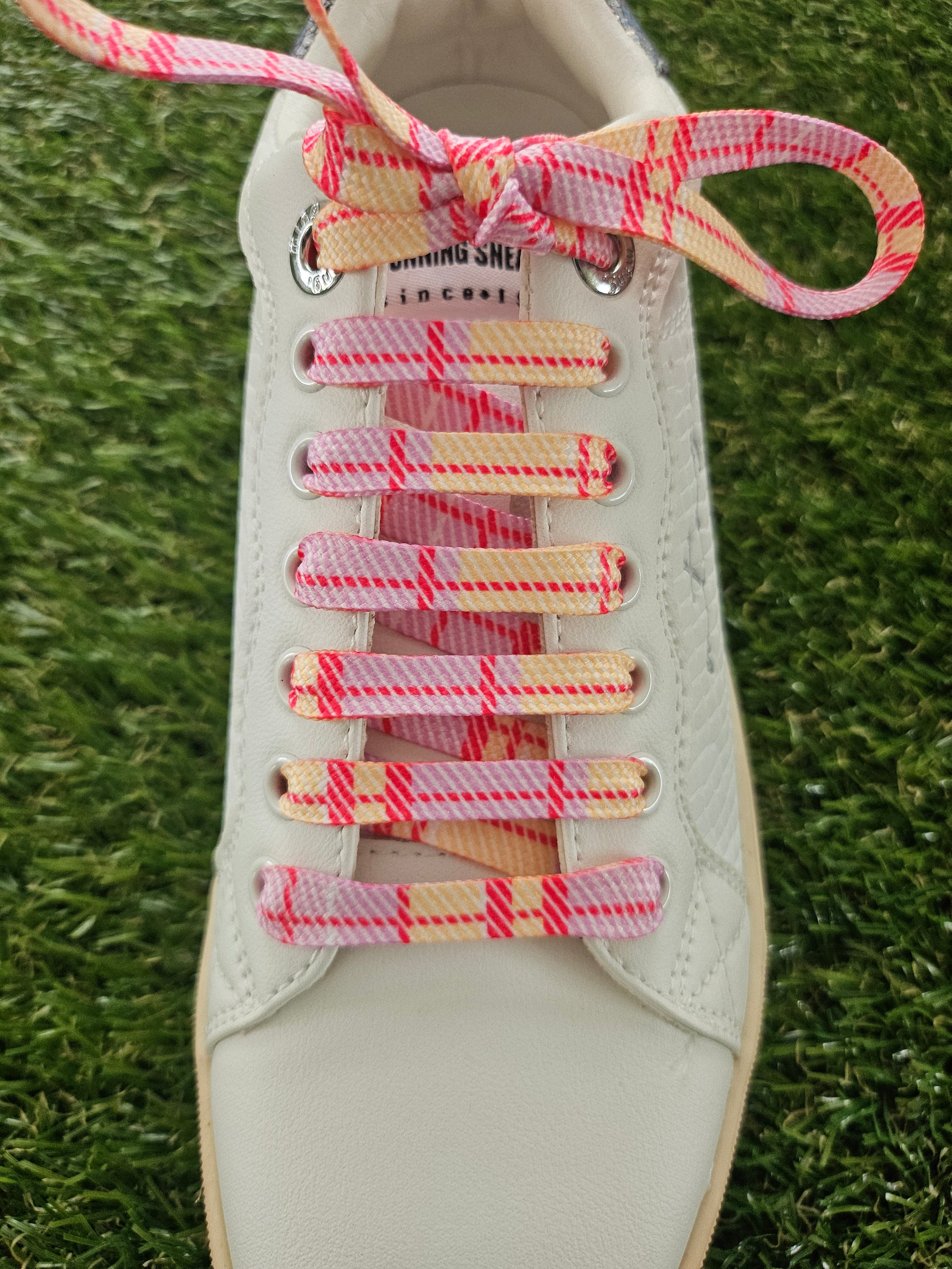 Close-up of a white sneaker with pink and yellow tartan checkered laces on grass.