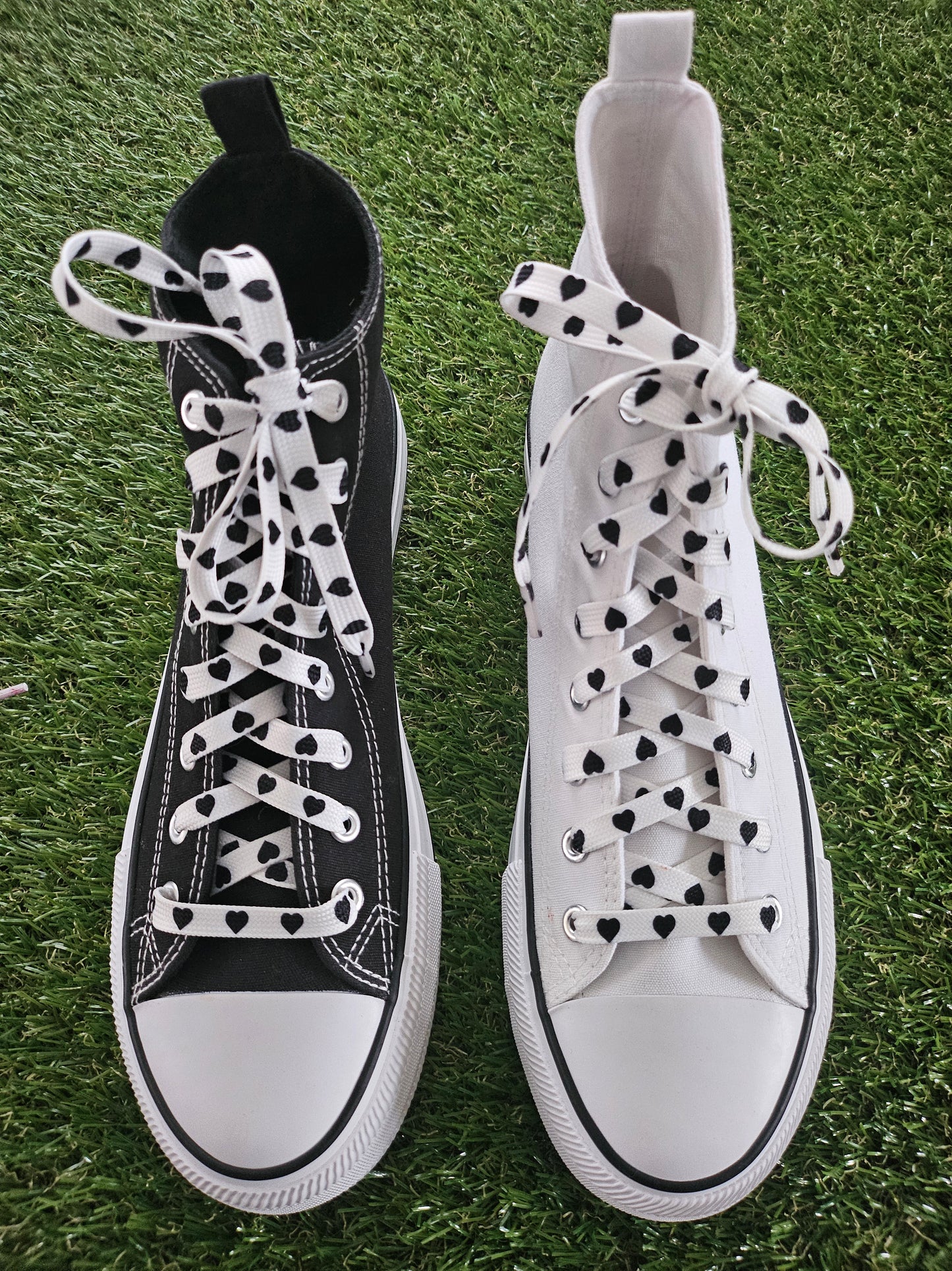 Pair of white and black high-top converse boots sneakers with baclk heart laces on grass.