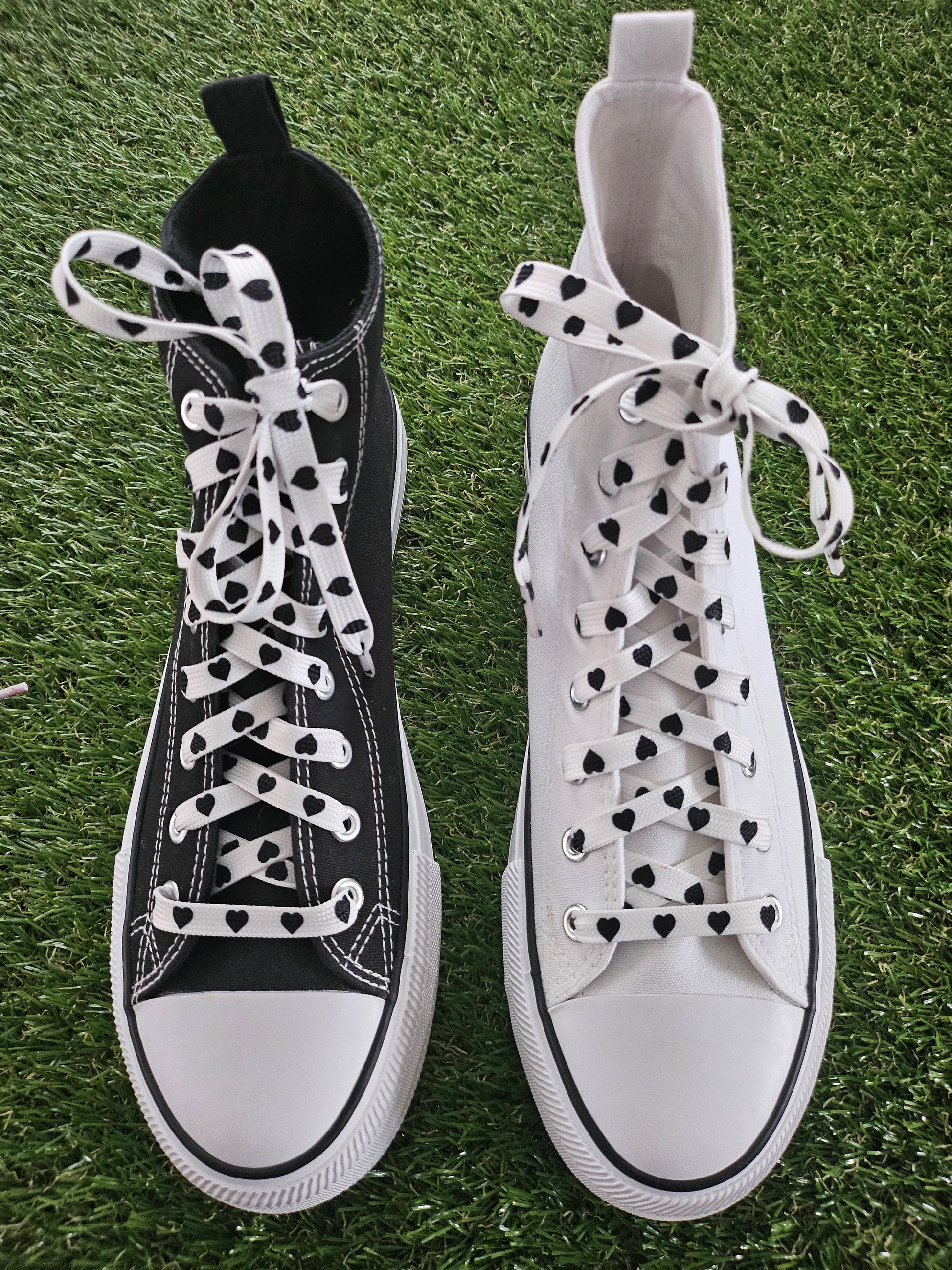 Pair of white and black high-top converse boots sneakers with baclk heart laces on grass.