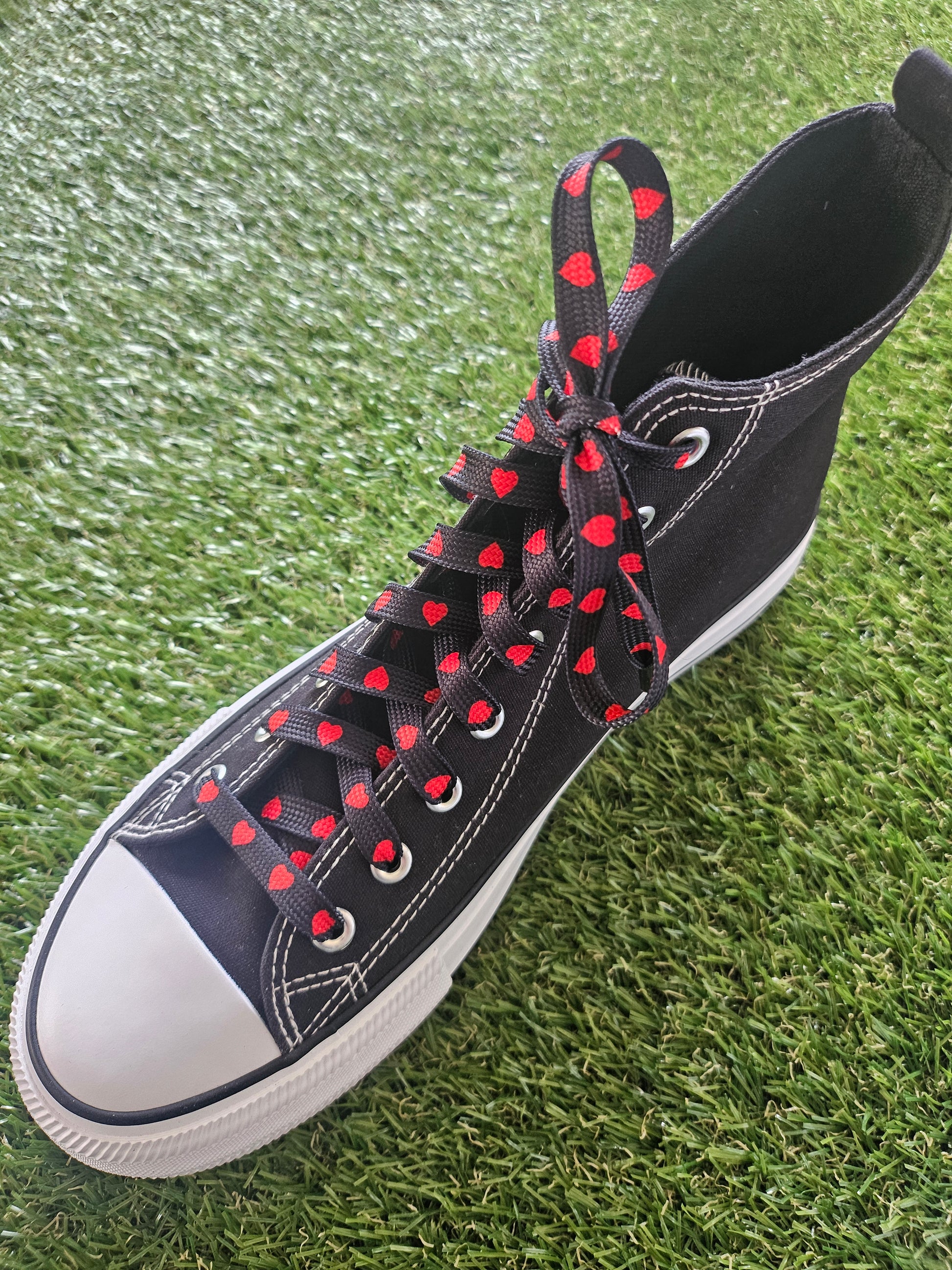 Black high-top sneaker with red heart patterns on laces against a grass background